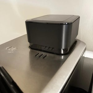 Puede incluir: Un electrodoméstico negro y rectangular con una parte superior brillante se encuentra sobre una superficie de metal cepillado. El electrodoméstico tiene un diseño elegante y moderno. La superficie metálica tiene tres ranuras paralelas y estrechas.