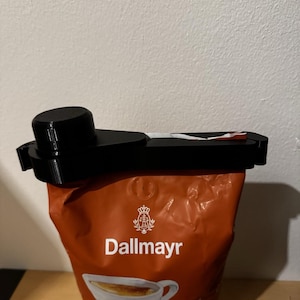 Puede incluir: Un clip de plástico negro está sujeto a una bolsa marrón de café Dallmayr Barista. La bolsa tiene un logotipo y texto blancos. El clip está diseñado para sellar la bolsa.
