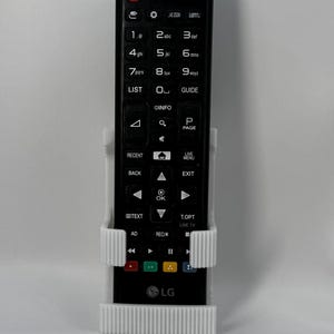 Puede incluir: Un mando a distancia de TV LG negro se sujeta en un soporte blanco impreso en 3D. El mando tiene numerosos botones, incluyendo teclas num&eacute;ricas, controles de navegaci&oacute;n y botones de funci&oacute;n. El logotipo de LG es visible en la parte inferior.