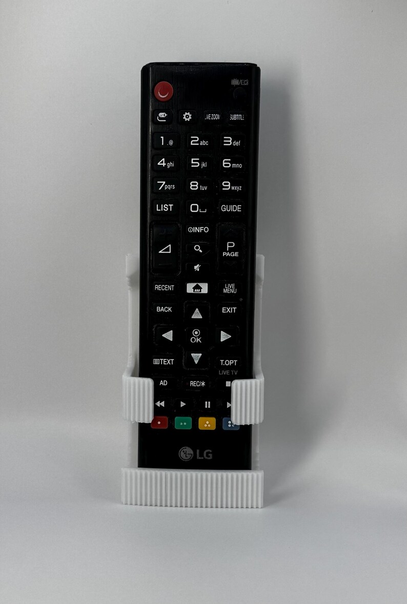 Puede incluir: Un mando a distancia de TV LG negro se sujeta en un soporte blanco impreso en 3D. El mando tiene numerosos botones, incluyendo teclas num&eacute;ricas, controles de navegaci&oacute;n y botones de funci&oacute;n. El logotipo de LG es visible en la parte inferior.