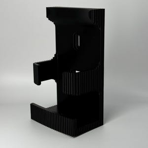 Puede incluir: Un soporte para tel&eacute;fono negro impreso en 3D con un dise&ntilde;o &uacute;nico en capas. El soporte presenta una textura acanalada en la base y los lados, y una ranura para que el tel&eacute;fono descanse. El soporte est&aacute; dise&ntilde;ado para sostener un tel&eacute;fono en posici&oacute;n vertical.