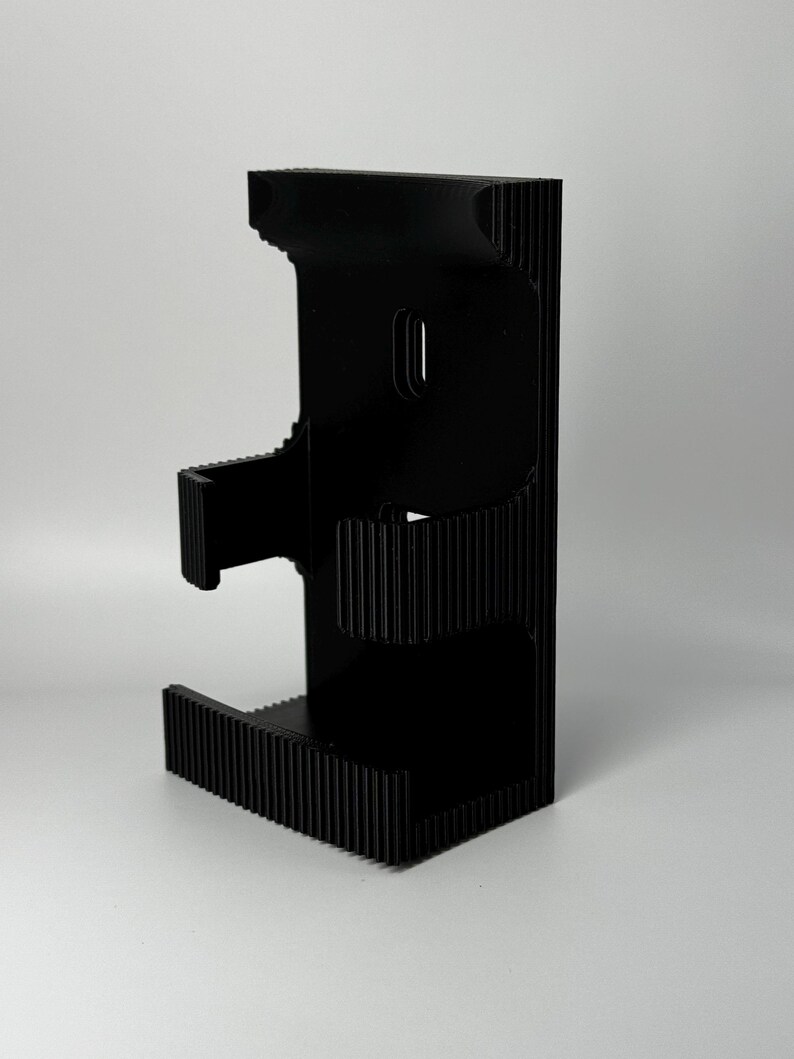 Puede incluir: Un soporte para tel&eacute;fono negro impreso en 3D con un dise&ntilde;o &uacute;nico en capas. El soporte presenta una textura acanalada en la base y los lados, y una ranura para que el tel&eacute;fono descanse. El soporte est&aacute; dise&ntilde;ado para sostener un tel&eacute;fono en posici&oacute;n vertical.