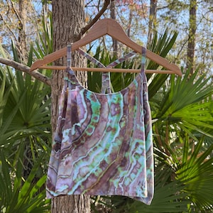 May include: A tie-dye tank top with a unique pattern of brown, green, and purple hues hangs on a wooden hanger. The tank top has a racerback design and is displayed outdoors, surrounded by trees and foliage.
