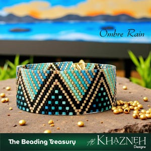 Op de afbeelding: Een kralenarmband met een ombre-ontwerp in tinten turquoise, blauw en zwart, geaccentueerd met gouden kralen. De armband heeft een geometrisch patroon en wordt gepresenteerd op een stenen oppervlak. De tekst "Ombre Rain" en "The Beading Treasury" zijn zichtbaar.