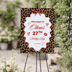 Puede incluir: Un letrero de bienvenida para una fiesta con un fondo de estampado de leopardo y un centro blanco festoneado. El letrero dice "Bienvenida a la fiesta de cumplea&ntilde;os n&uacute;mero 27 de Olivia" en escritura roja, con corazones, estrellas y un lazo decorativos.