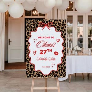 Puede incluir: Un letrero de bienvenida para una fiesta con un fondo de estampado de leopardo y un marco blanco festoneado. El letrero dice "Bienvenida a la fiesta de cumplea&ntilde;os n&uacute;mero 27 de Olivia" en escritura roja e incluye un lazo rojo, corazones y una marca de beso. Tambi&eacute;n se muestra la fecha 11.22.2025.