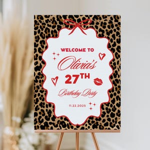 Puede incluir: Un letrero de bienvenida para una fiesta de cumplea&ntilde;os con un fondo de estampado de leopardo y un marco blanco festoneado. El letrero dice "Welcome to Olivia's 27th Birthday Party" en escritura roja, con corazones, estrellas y un gr&aacute;fico de beso. La fecha es 11.22.2025.