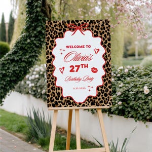Puede incluir: Un caballete de madera exhibe un letrero con un borde de estampado de leopardo. El letrero dice "Bienvenida a la fiesta de cumplea&ntilde;os n&uacute;mero 27 de Olivia" con corazones, estrellas y un beso. Tambi&eacute;n se muestra la fecha 11.22.2025.