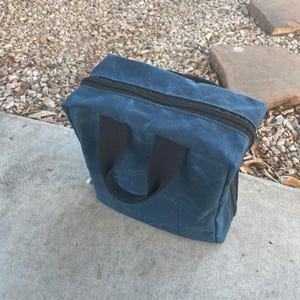 May include: A blue canvas tote bag with a black zipper and black handles. The bag is sitting on a concrete surface. The bag is a square shape and appears to be empty.