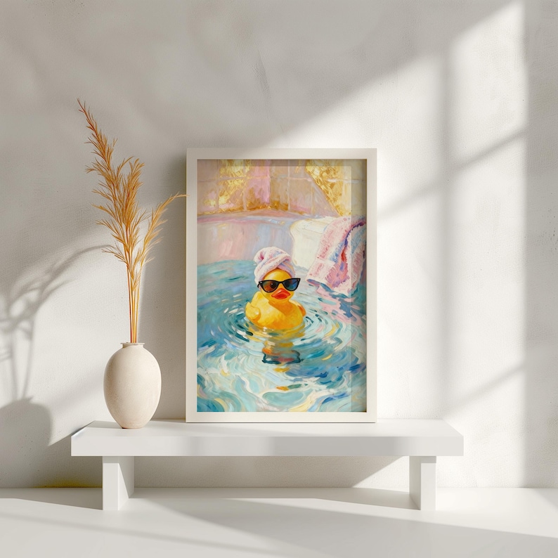 May include: A framed art print featuring a yellow rubber duck wearing sunglasses and a pink towel on its head, relaxing in a bathtub. The artwork has a painterly style with blue and pink water and a white frame.
