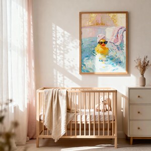 May include: A framed art print depicts a yellow rubber duck wearing sunglasses and a towel on its head, relaxing in a bathtub. The artwork is in a light-filled room with a wooden crib, a beige blanket, and a white dresser.