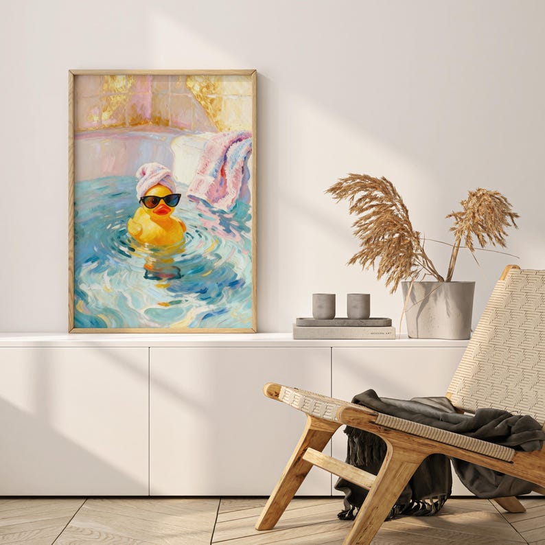 May include: A framed art print depicts a yellow rubber duck wearing sunglasses and a pink towel on its head, relaxing in a blue and white bath. The artwork has a light wood frame and is displayed on a white cabinet. A wooden chair and decorative plants are in the background.