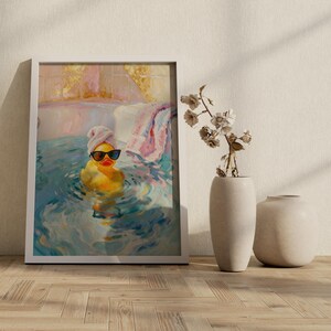 May include: Framed art print of a yellow rubber duck wearing sunglasses and a towel on its head, relaxing in a bathtub. The painting has a colorful, impressionistic style with blue, pink, and yellow hues. A towel hangs on the edge of the tub.