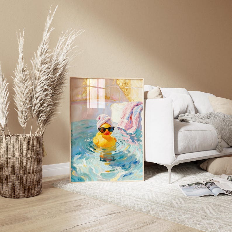 May include: A framed art print depicts a yellow rubber duck wearing sunglasses and a towel on its head, relaxing in blue water. A pink and white towel hangs nearby. The artwork is displayed in a room with a white couch and a woven basket.