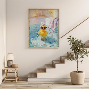 May include: A framed painting depicts a yellow rubber duck wearing sunglasses and a towel on its head, floating in blue water. The artwork has a light wood frame and is displayed on a white wall. A small table with a lamp and a plant are in the foreground.