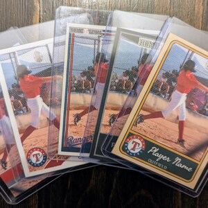 May include: Baseball trading cards featuring a player in a red uniform swinging a bat. The cards are in protective sleeves, with the Texas Rangers logo visible. The cards are on a wooden surface.