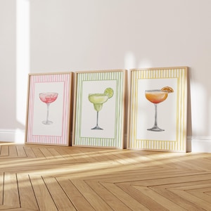 May include: Three framed watercolor prints of cocktails. The first features a pink drink in a coupe glass, the second a margarita with a lime, and the third an orange cocktail. Each print has a striped border and a wooden frame.