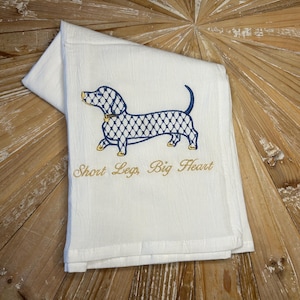 May include: White folded kitchen towel with an embroidered dachshund design in navy blue and gold. The dachshund has a lattice pattern. The text "Short Leg, Big Heart" is embroidered below the dog.