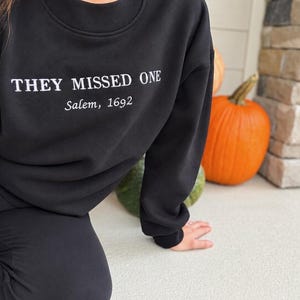 以下が含まれることがあります： 胸元に「THEY MISSED ONE」という太字の白い大文字テキストがプリントされた黒いクルーネックのスウェットシャツ。そのすぐ下に、より小さな斜体のテキストで「Salem, 1692」と中央揃えで記載されています。