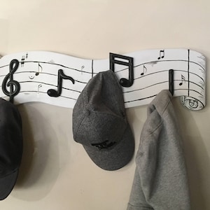 Puede incluir: Un perchero de pared de madera blanca con forma de pentagrama musical, adornado con notas musicales negras y ganchos. Tres artículos cuelgan de los ganchos: una gorra negra, una gorra gris y una sudadera gris. El perchero tiene un diseño en forma de pergamino.