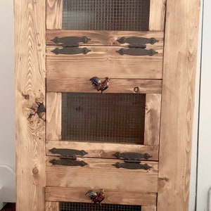May include: A rustic wooden cabinet with four sections, each featuring a wire mesh panel and decorative rooster-shaped handles. The cabinet has a natural wood finish and metal hinges, ideal for storage.