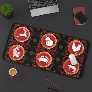 Puede incluir: Alfombrilla de ratón rectangular negra con un patrón repetitivo de diseños geométricos dorados y rojos. La alfombrilla presenta seis círculos rojos con ilustraciones blancas de un ciervo, una calabaza, un pez, un cangrejo, un gallo y un camarón. Un ratón de ordenador descansa sobre la alfombrilla.