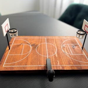 Könnte beinhalten: Ein Miniatur-Basketballspiel aus Holz. Das Spielfeld ist braun mit weißen Linien, und an jedem Ende befinden sich zwei kleine Basketballkörbe. Ein schwarzer Griff befindet sich in der Mitte des Spielfelds.