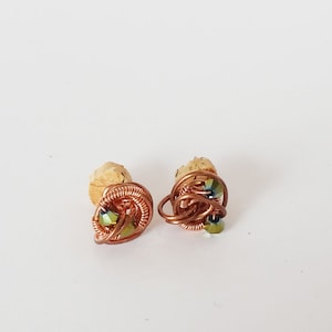 May include: Two handmade copper wire earrings with a woven design, each featuring a small, iridescent green bead. The earrings are displayed on a light-colored surface, showcasing their intricate wirework and colorful accents.