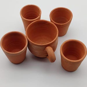 Peut inclure: Ensemble de poterie en terre cuite non émaillée. Il comprend une grande tasse avec une anse et quatre petites tasses. Les objets sont de couleur brun-orangé chaud et semblent faits à la main.