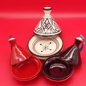 Peut inclure: Ensemble de tagines marocains, comprenant un grand couvercle blanc et noir à motifs, une assiette crème avec un motif central et deux tagines rouges et noirs plus petits. Le fond est rouge uni.