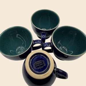 May include: Four navy blue Denby England mugs with teal interiors. Each mug has a handle and a speckled finish. The bottom of one mug displays the Denby England logo and care instructions. The mugs are microwave, freezer, and oven safe.