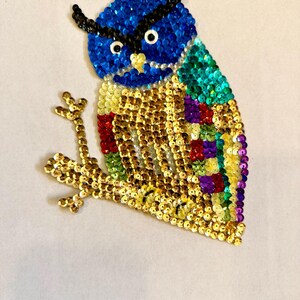 May include: A colorful sequined owl applique. The owl features a blue head, gold body, and accents of green, red, and purple. It is perched on a gold branch, creating a decorative and eye-catching design.