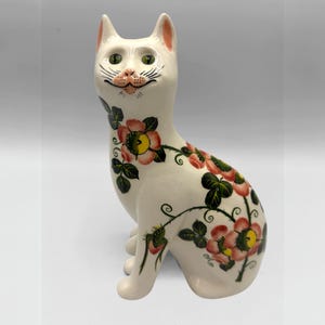 May include: A white ceramic cat figurine with green eyes, pink ears, and a pink nose. The cat is decorated with pink and yellow floral patterns and green leaves. The cat is sitting upright with its tail curled.
