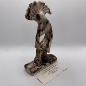May include: A carved stone sculpture of a Native American figure with a headdress, in shades of beige and brown. The figure is bent over, with arms at its sides. A certificate of authenticity is attached.