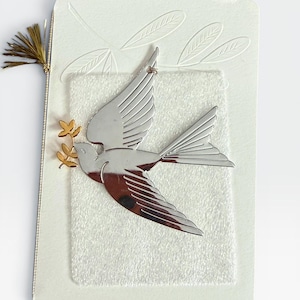 May include: A silver-toned metal bird ornament with a gold-toned olive branch in its beak, attached to a rectangular off-white card. The card features an embossed leaf design and a small gold tassel at the top left corner, with a textured white inlay.
