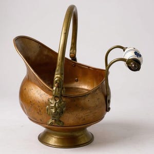 May include: Antique copper coal scuttle with a brass handle and decorative accents. The scuttle features a pedestal base and a small handle with a white and blue floral ceramic knob. A vintage piece for home decor.