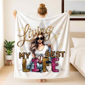 May include: White blanket with the words "Living My Best Life" in gold and pink leopard print. The blanket features an illustration of a woman wearing sunglasses and a denim jacket.