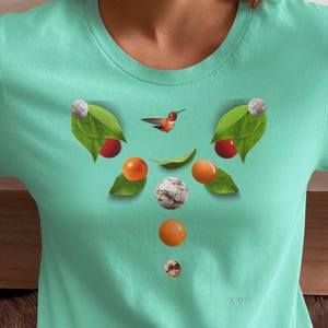 May include: A light teal t-shirt featuring a nature-inspired design. The graphic includes a hummingbird, leaves, and various fruits in shades of red, orange, and white. The shirt has short sleeves and a crew neck.
