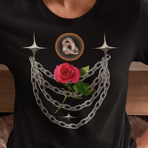 May include: Black t-shirt with a graphic design featuring a red rose, silver chains, and a circular knot-like element. The design is printed on a crew neck, short-sleeved t-shirt. The style combines floral and chain elements for a unique look.