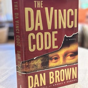 May include: A hardcover book titled The Da Vinci Code by Dan Brown. The cover is deep red with gold lettering and features a torn section revealing a portion of the Mona Lisa painting and handwritten script. The spine also displays the title and author.