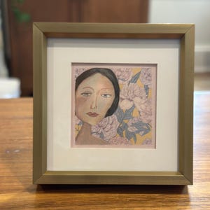 May include: Framed artwork depicting a woman's portrait with a floral backdrop. The artwork is in a gold-coloured frame with a white mount. The woman's face is painted with soft colours, and the background has pink and yellow flowers.