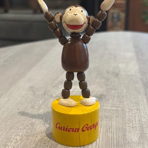 May include: A wooden Curious George monkey figurine with brown limbs made of beads, a cream-colored face, and red mouth. The monkey stands with arms raised on a cylindrical yellow base featuring the red text Curious George printed on the front.