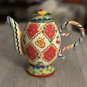 May include: A fabric teapot ornament featuring a red and cream diamond pattern with cherry and flower motifs. The spout and handle have a multi-colored striped design. A small yellow knob sits atop the lid, which has a green floral print.