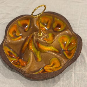 May include: A vintage ceramic serving dish with a brown exterior and a divided interior featuring orange and yellow accents. It has a gold-colored handle for easy carrying. The dish is designed for serving snacks or appetizers.