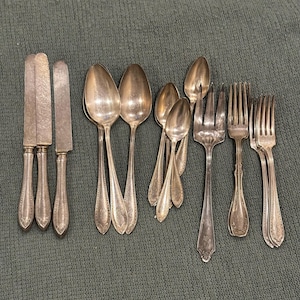 May include: A collection of antique silverware, including knives, spoons, and forks, arranged on a textured green surface. The silverware has ornate detailing on the handles and a tarnished silver finish, suggesting age and use. The arrangement showcases a variety of dining utensils.