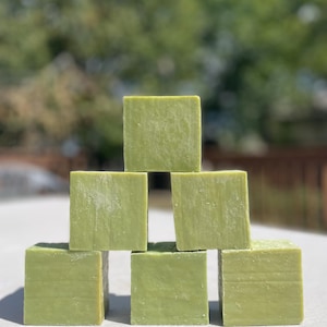 May include: A pyramid of seven square, light green soap bars. The bars are stacked on a white surface, with a blurred green background of trees and foliage. The soap bars have a textured appearance.