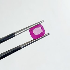 Op de afbeelding: Een levendige, kussenvormig geslepen robijn, vastgehouden door een zilveren pincet. De robijn vertoont een rijke, dieproze tint met een gefacetteerd oppervlak, dat licht reflecteert. De pincet is geplaatst tegen een effen witte achtergrond.