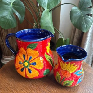 May include: Two ceramic jugs with a vibrant floral design. The jugs are red with yellow, orange, and blue flower patterns and green leaves. The interiors and handles are blue. The jugs are on a wooden surface.