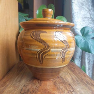 May include: A ceramic jar with a lid, featuring a warm brown and orange color scheme. The jar has a rounded shape with decorative dark brown wave-like patterns. The lid has a small knob. The jar is sitting on a wooden surface.
