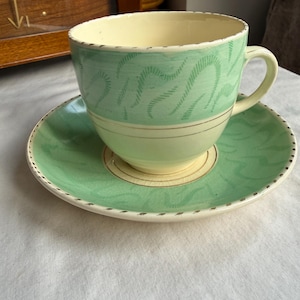 May include: A vintage teacup and saucer set. The cup and saucer are primarily light green with a cream-colored interior and base. The cup features a wavy pattern, and the saucer has a matching design. Gold trim accents the edges.